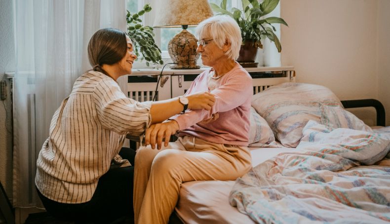 A caregiver helps an older woman get up from her bed.