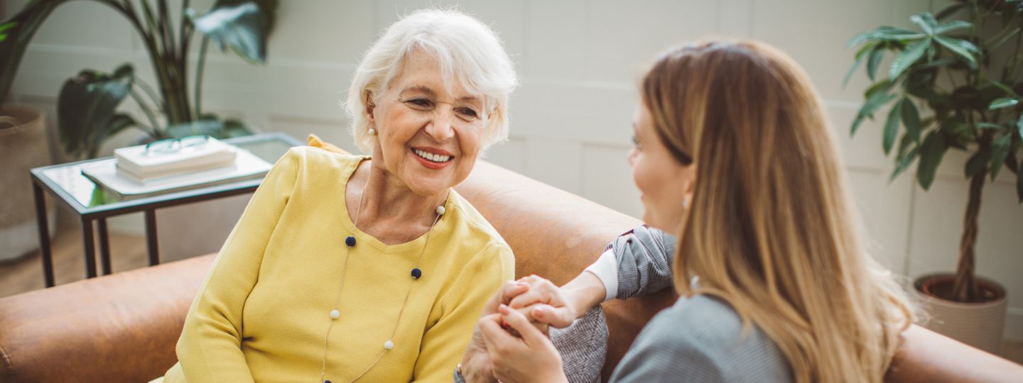 A caregiver and older adult chat while sitting together on a couch.