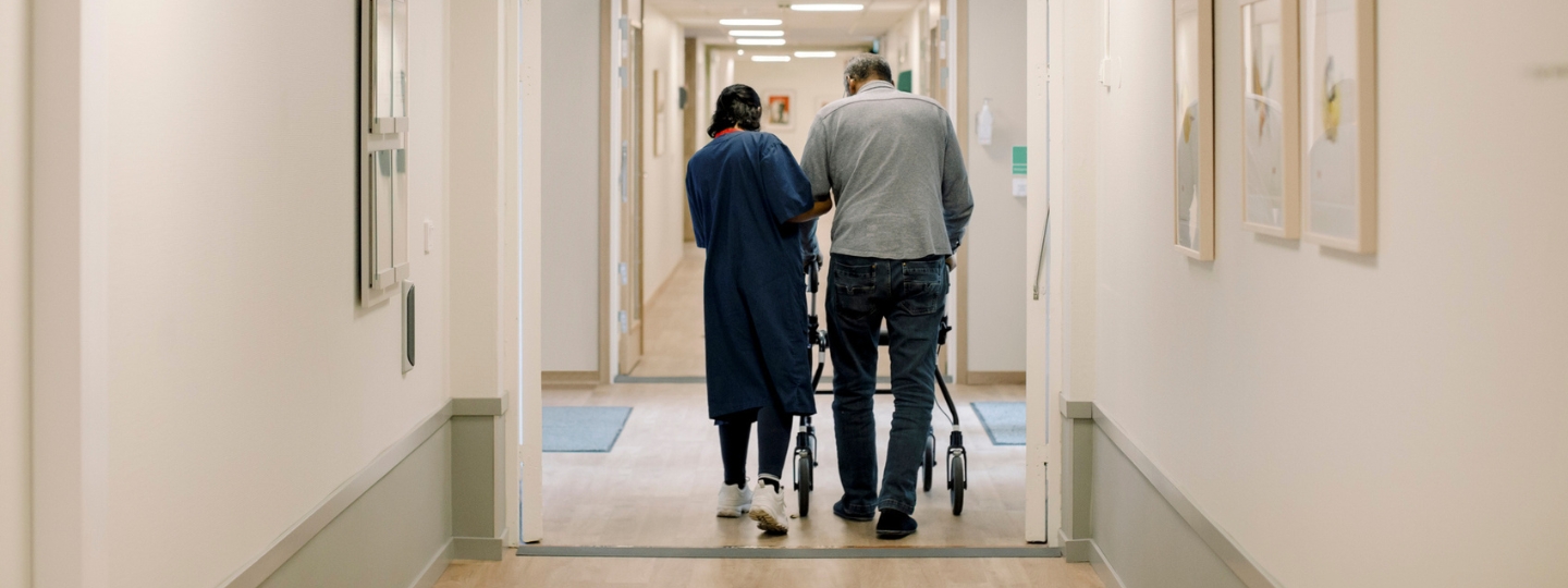 A caregiver helps an older adult walk down a hallway with their walkers.