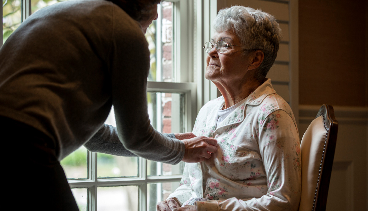 Woman wearing glasses sitting in chair receiving assistance buttoning her shirt by nursing home professional